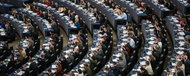 Wide-angle view of the European Parliament in session, with numerous Members of the European Parliament (MEPs) seated in curved rows, many raising their hands to vote. The chamber is full, and desks are equipped with documents, microphones, and electronic voting systems.