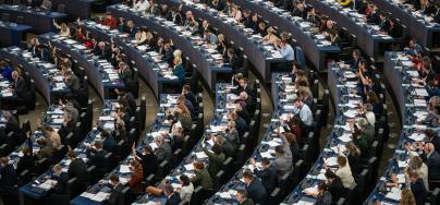 Wide-angle view of the European Parliament in session, with numerous Members of the European Parliament (MEPs) seated in curved rows, many raising their hands to vote. The chamber is full, and desks are equipped with documents, microphones, and electronic voting systems.