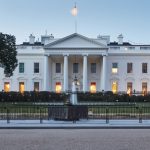 A clear, symmetrical shot of the White House in Washington, D.C., taken at dawn. The soft morning light contrasts with the warm glow emanating from the windows of this historic building. The U.S. flag flies at the top. — Photo by jonkempner
