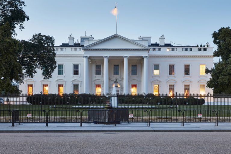 A clear, symmetrical shot of the White House in Washington, D.C., taken at dawn. The soft morning light contrasts with the warm glow emanating from the windows of this historic building. The U.S. flag flies at the top. — Photo by jonkempner