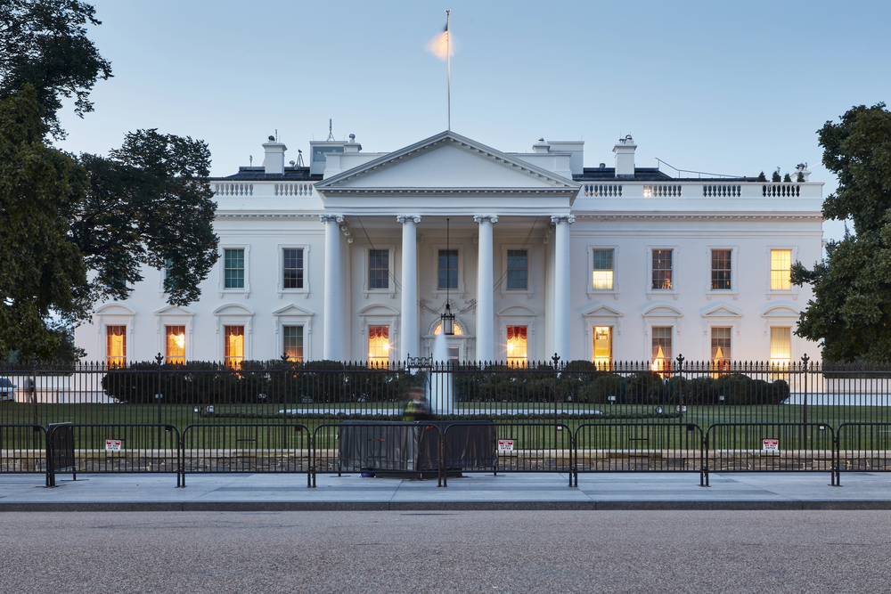 A clear, symmetrical shot of the White House in Washington, D.C., taken at dawn. The soft morning light contrasts with the warm glow emanating from the windows of this historic building. The U.S. flag flies at the top. — Photo by jonkempner